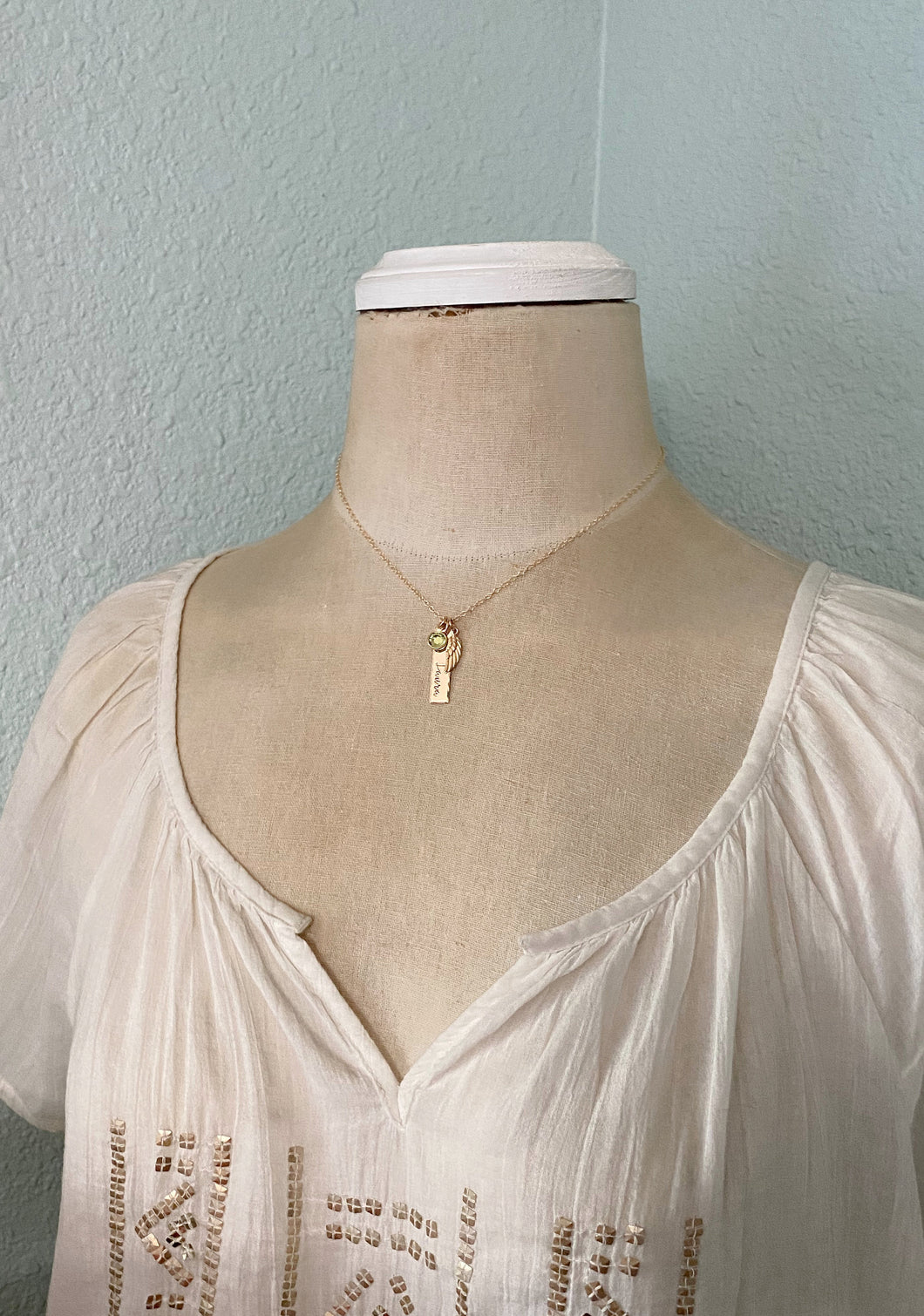 Gold tone necklace with birthstone crystal, bar with name and angel wing charm shown on mannequin